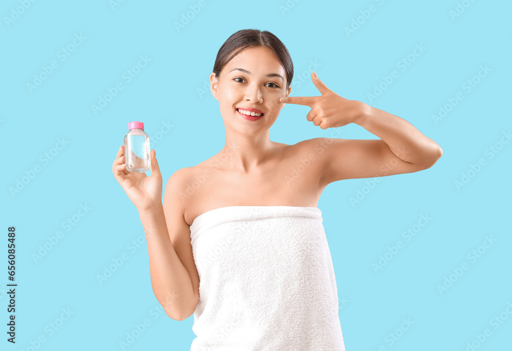 Young Asian woman with bottle of micellar water pointing at her face on blue background