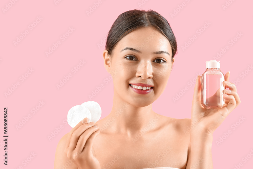 Young Asian woman with bottle of micellar water and cotton pads on pink background, closeup