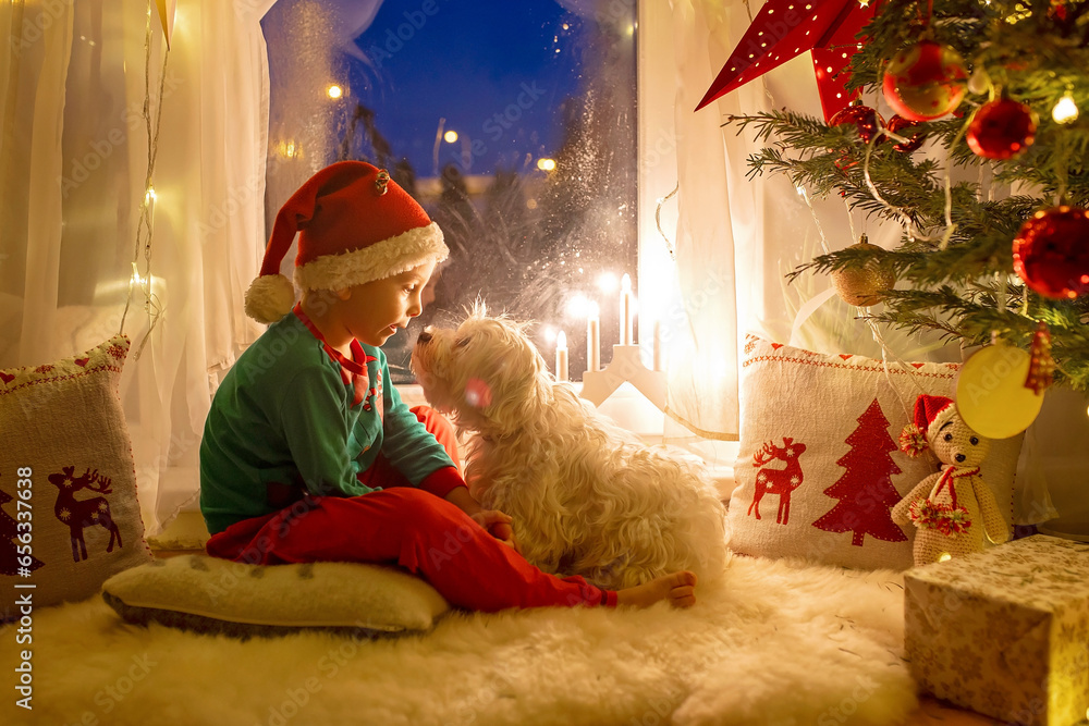 Toddler child, cute blond boy, sitting on the window in pajama, looking out for Satna Claus, Christmas lights around him