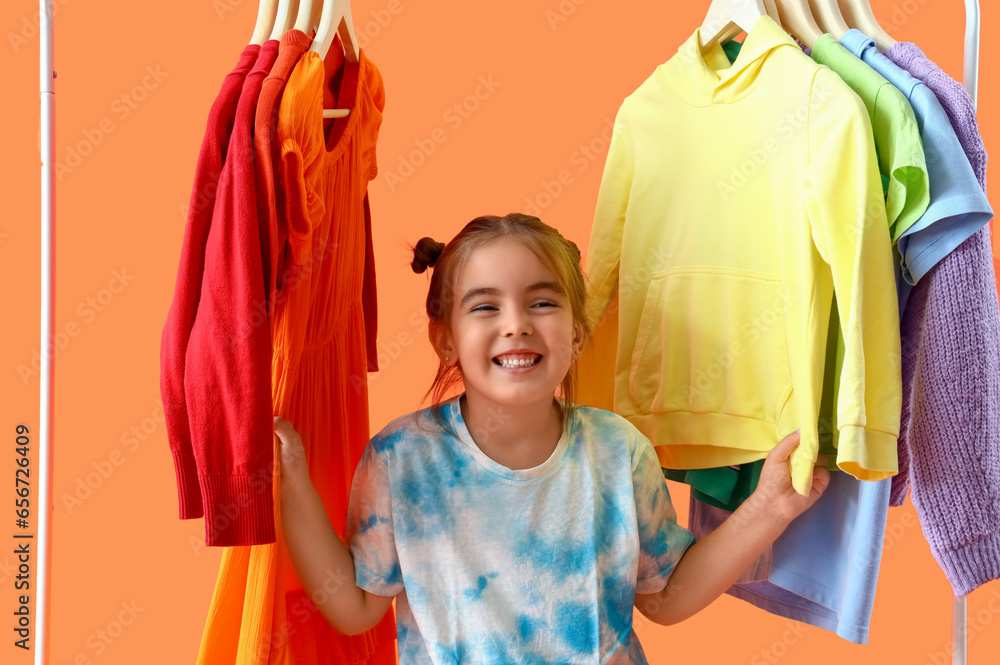 Cute little girl and rack with clothes on orange background