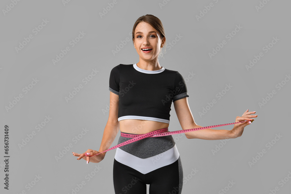 Beautiful young woman measuring her waist on grey background. Weight loss concept