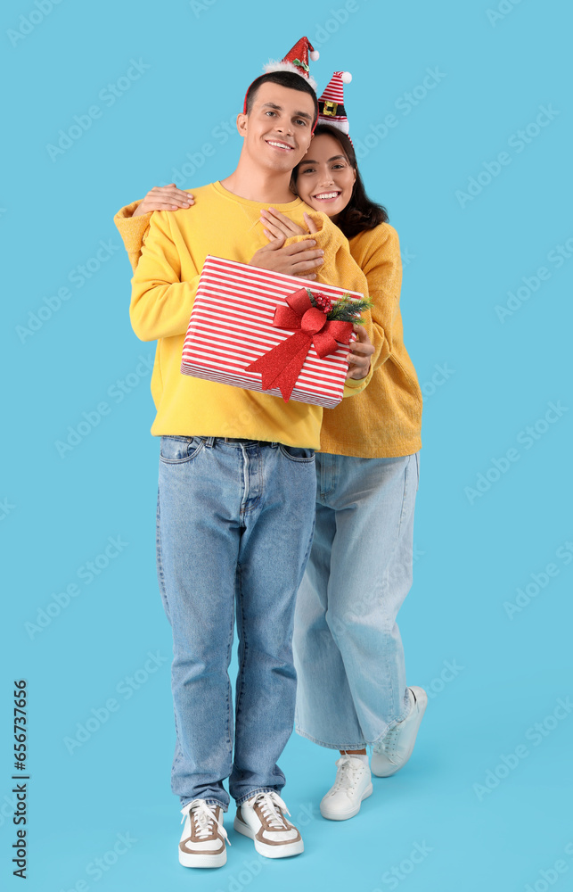 Happy young couple in Christmas hats with gift box on blue background