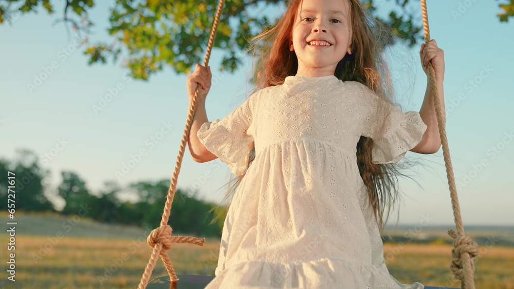 Happy little girl swings on swing in park under tree at sunset. Baby swing, kid girl smile in flight. Child plays on wooden swing, dreams of flying. Concept of family happiness, dreams, entertainment