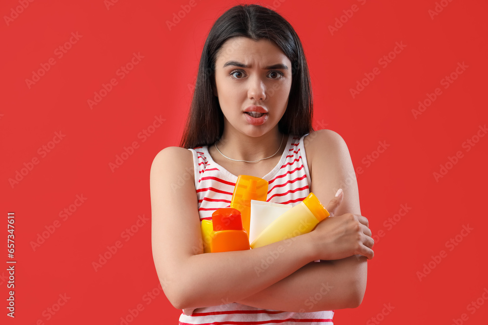 Worried young woman with bottles of sunscreen cream on red background