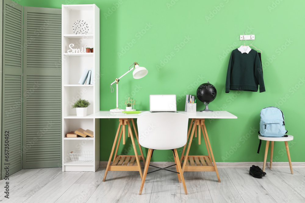 Workplace with desk, shelving unit, laptop computer, backpack and stylish school uniform hanging on green wall