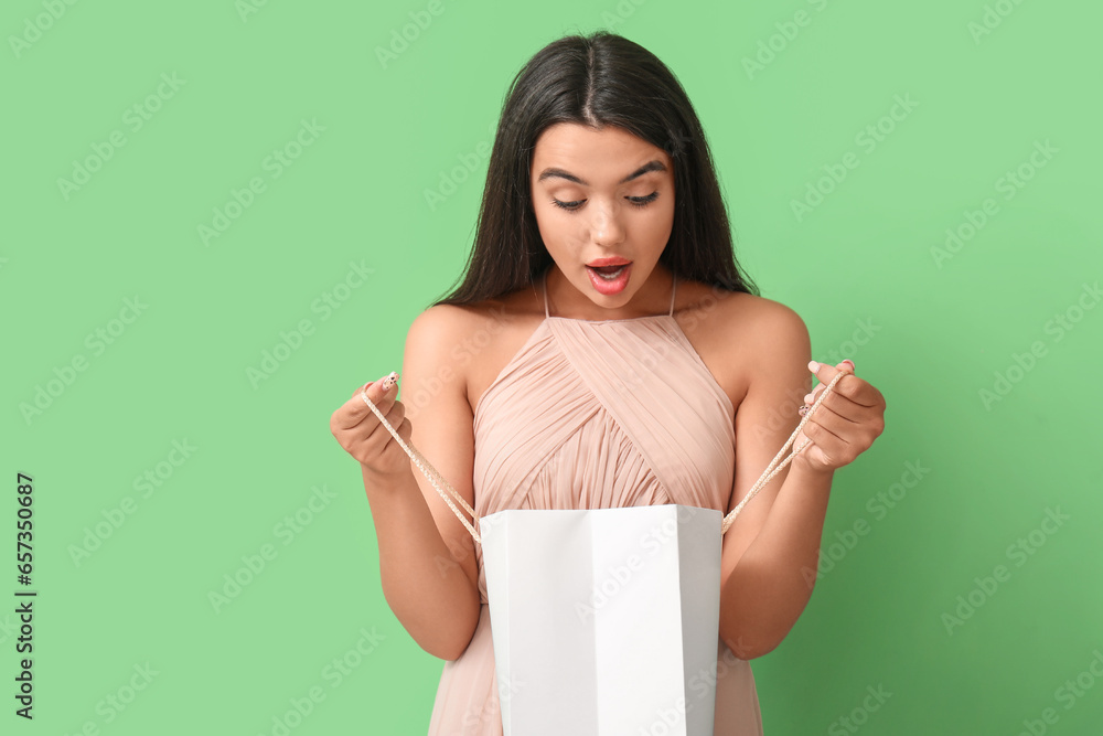 Shocked beautiful young woman opening shopping bag on green background