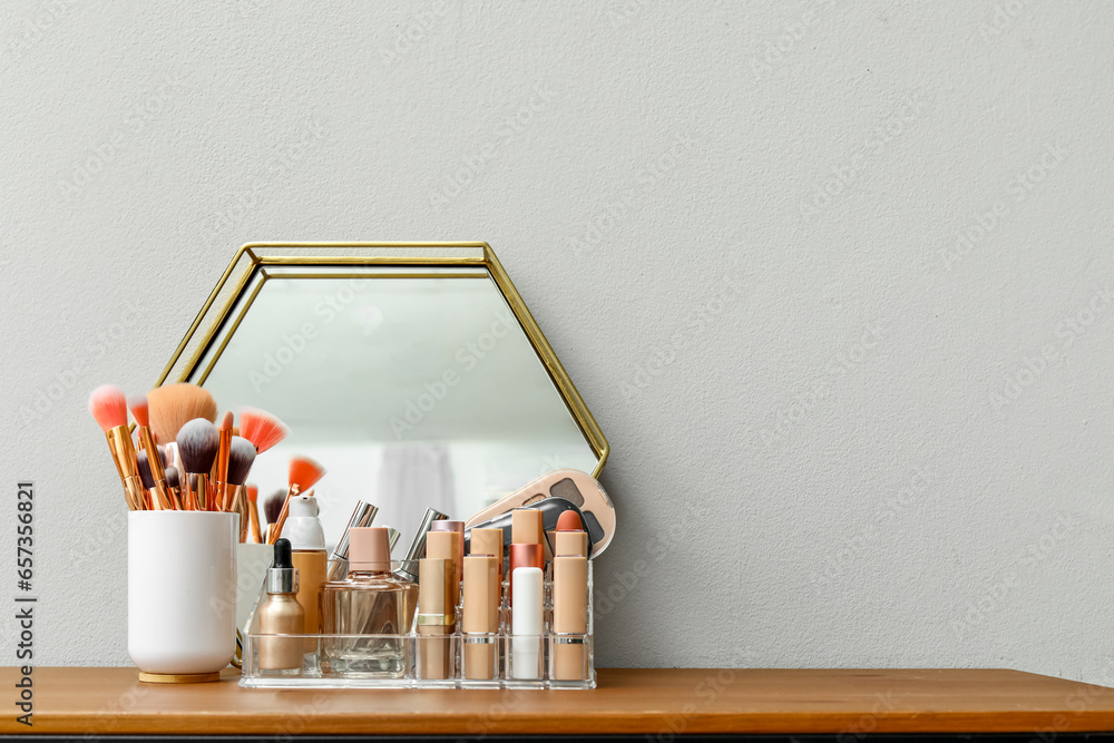 Holder with different makeup products and mirror on dressing table near light wall in room