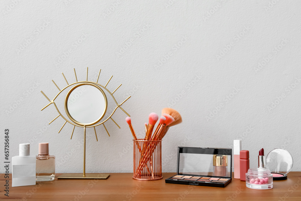 Different makeup products and mirror on dressing table near light wall in room