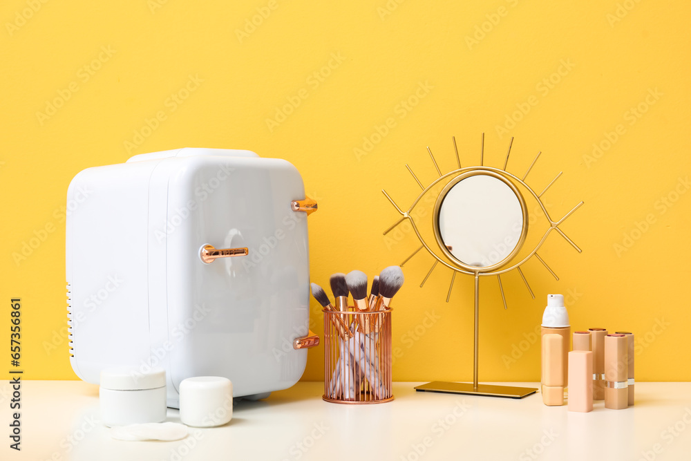 Portable fridge with different makeup products and mirror on dressing table near orange wall in room