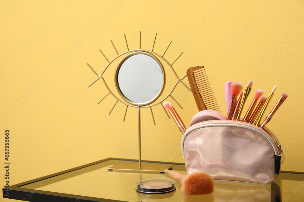 Cosmetic bag with different makeup products and mirror on dressing table near beige wall in room