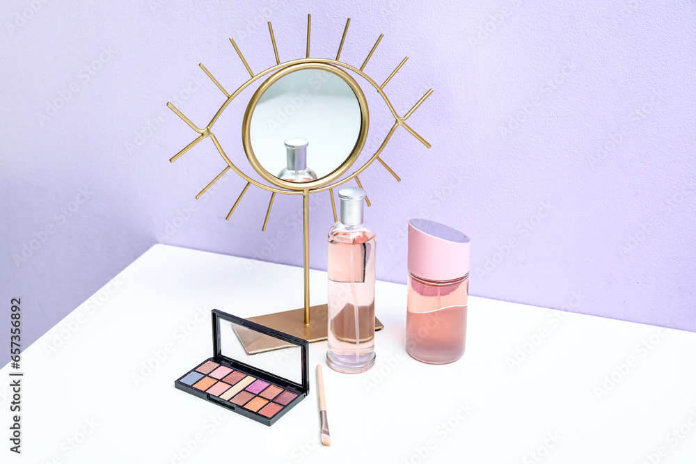 Bottles of perfume, eyeshadows, brush and mirror on dressing table near purple wall in room