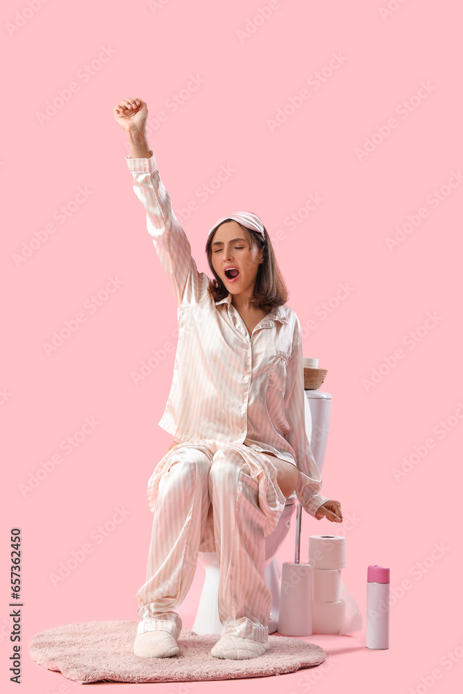 Sleepy young woman in pajamas sitting on toilet bowl against pink background