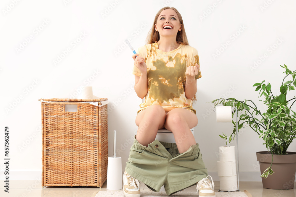 Happy young woman with pregnancy test sitting on toilet bowl in restroom