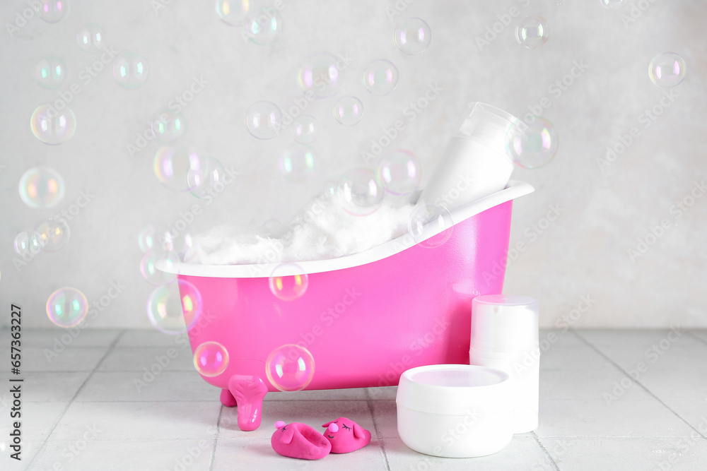 Small bathtub with cotton wool, cosmetic products and soap bubbles on light tile table