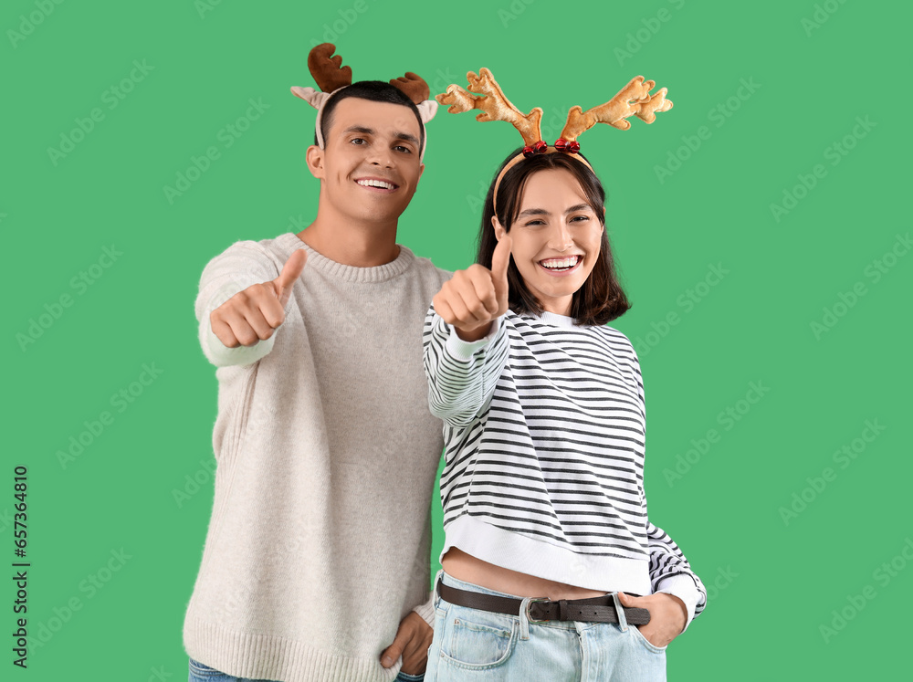 Happy young couple in Christmas reindeer horns showing thumbs-up gesture on green background