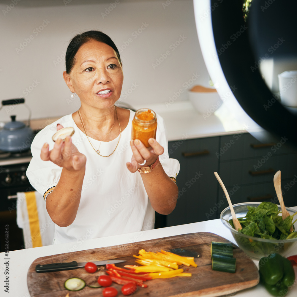 Asian woman, vegetables and live streaming for cooking with bottle, healthy food and blog on social media in kitchen. Chef, mature influencer and content creation, class and broadcast online tutorial