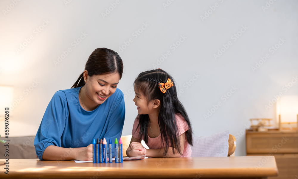 A young family spends their free time together in the living room at home. Mother and little daughter draw pictures with crayons on paper, smiling happy.