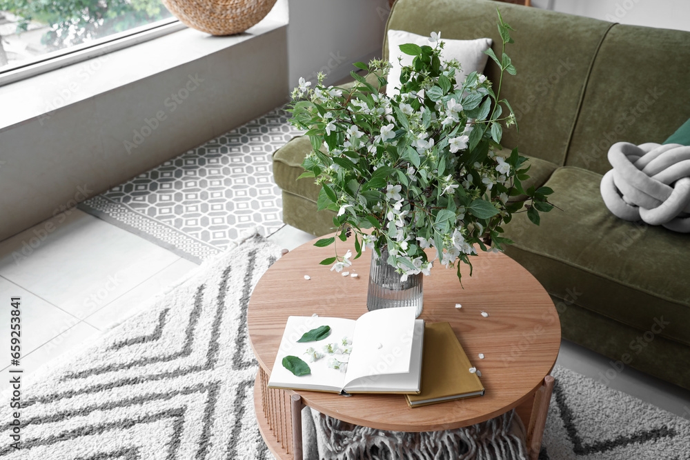 Vase with blooming jasmine flowers and books on wooden coffee table in living room