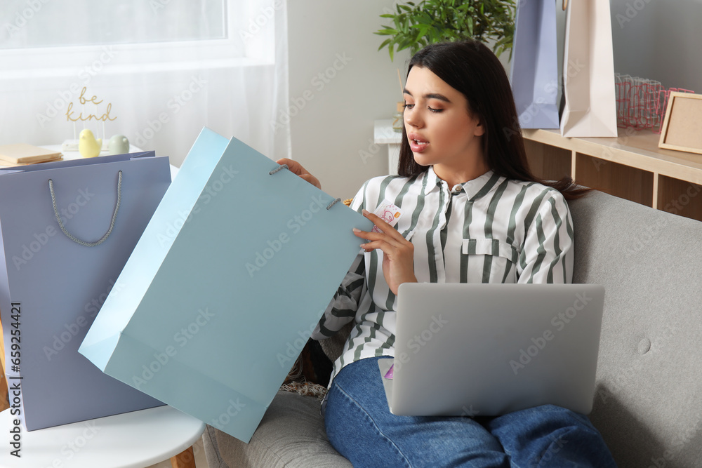 Young woman with gift card, shopping bag and laptop at home