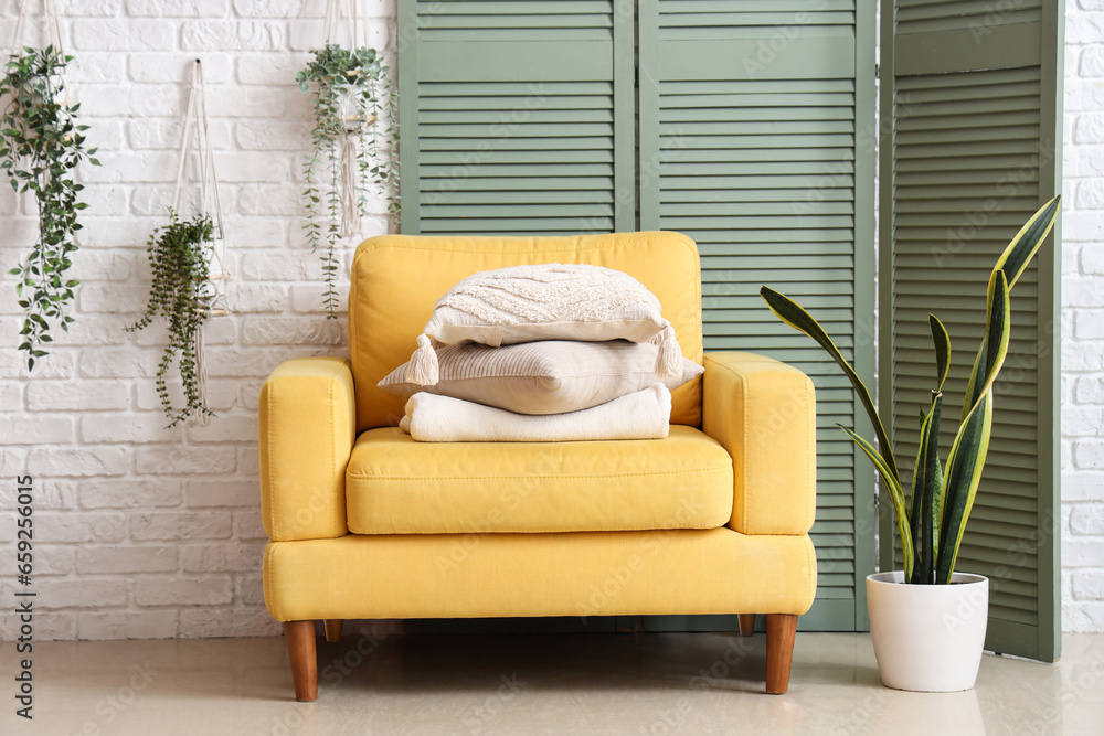 Yellow armchair with cushions and green folding screen near white brick wall