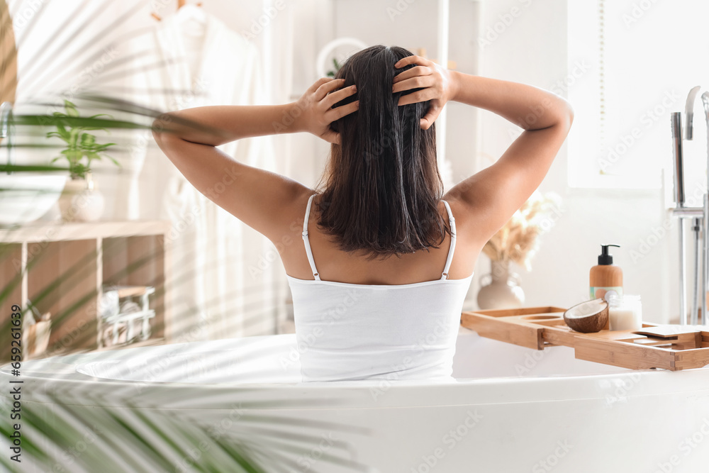 Pretty young woman applying coconut oil on her hair in bathroom, back view