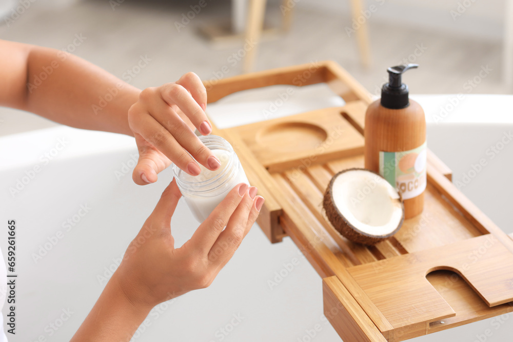 Beautiful young woman applying coconut oil on her body in bathroom, closeup