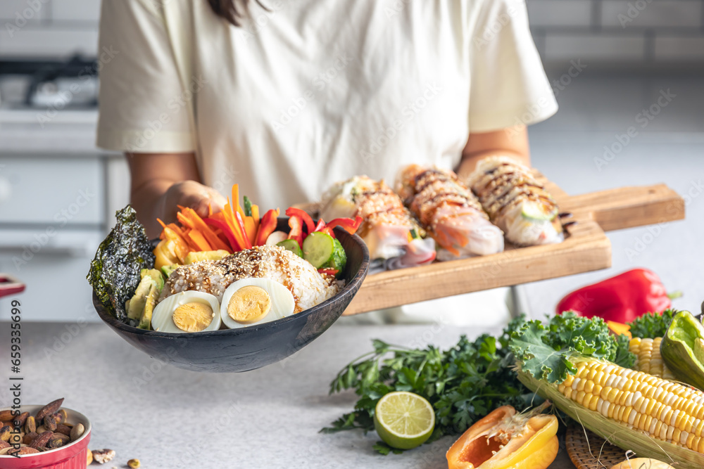 Bowl with vegetables and rice and spring rolls in female hands in the kitchen.