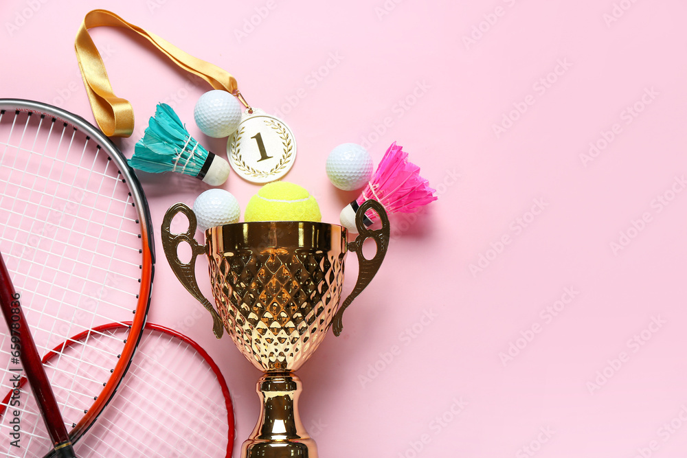 Gold cup with first place medal and sports equipment on pink background