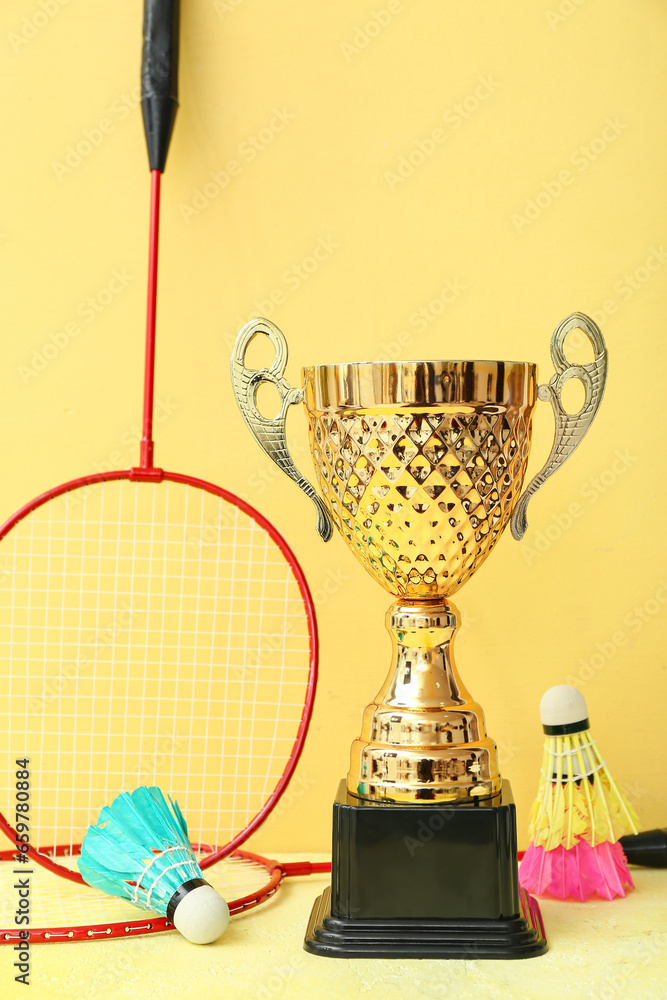 Gold cup with badminton rackets and shuttlecocks on yellow background