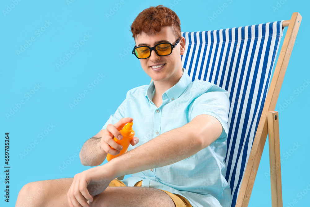 Young man applying sunscreen cream in deck chair on blue background