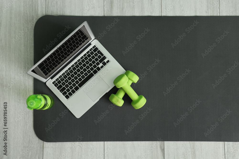 Laptop with dumbbells and bottle of water on fitness mat in gym, top view