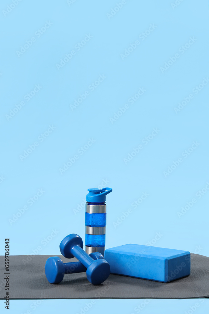 Fitness mat with bottle of water, block and dumbbells on blue background