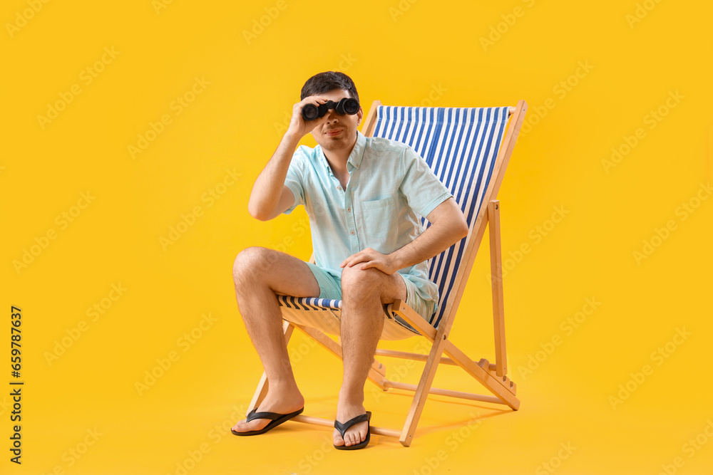 Young man with binoculars sitting in deck chair on yellow background