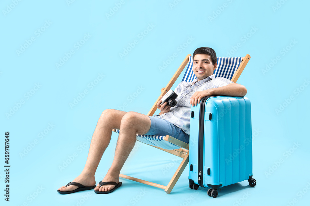 Young man with photo camera and suitcase sitting in deck chair on blue background