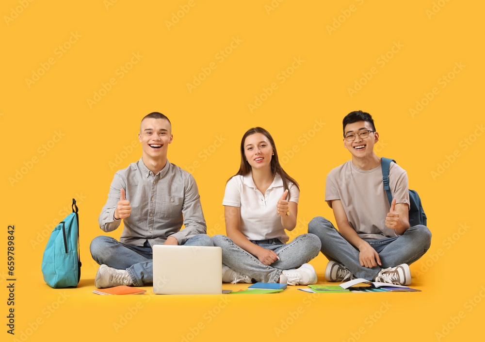 Happy students with notebooks and laptop sitting on yellow background and showing thumbs-up gesture