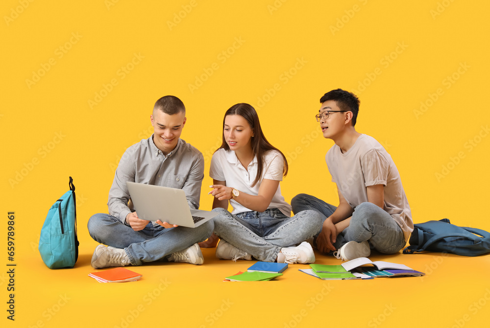 Happy students with backpacks, notebooks and laptop sitting on yellow background