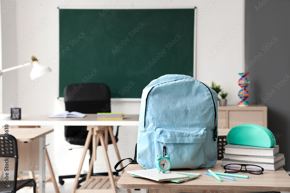 Blue school backpack with stationery, eyeglasses and alarm clock on desk in classroom