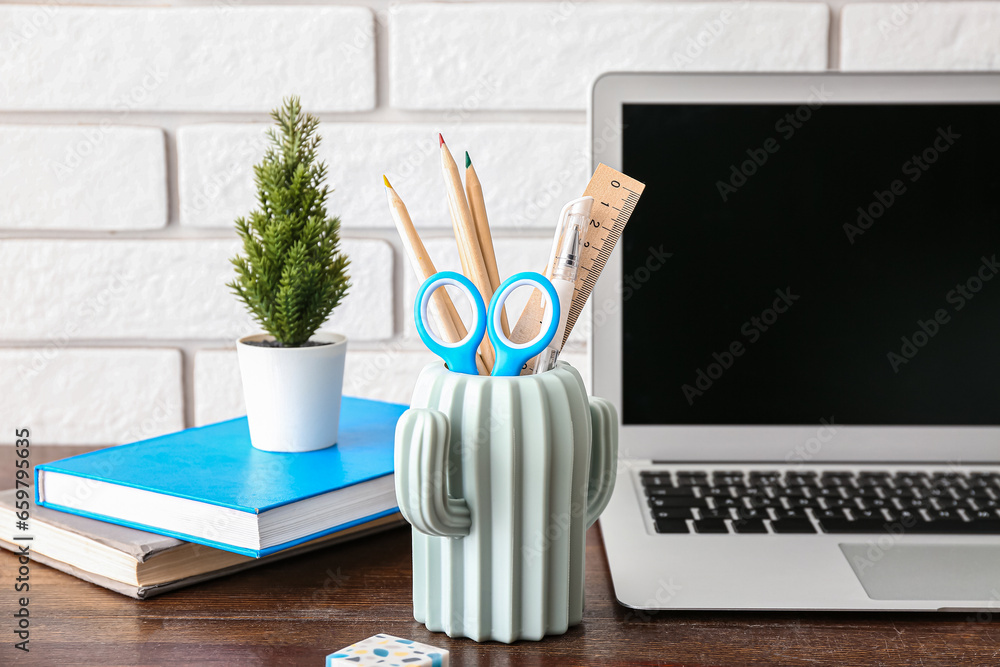 Modern laptop, houseplant and holder with different stationery on wooden desk, closeup