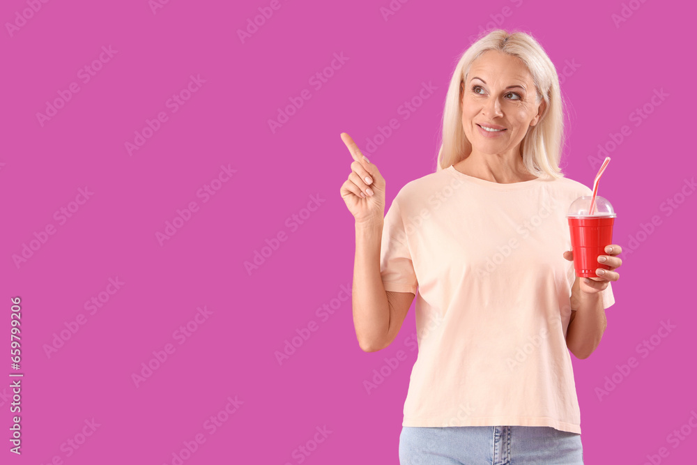 Mature woman with cup of soda pointing at something on purple background