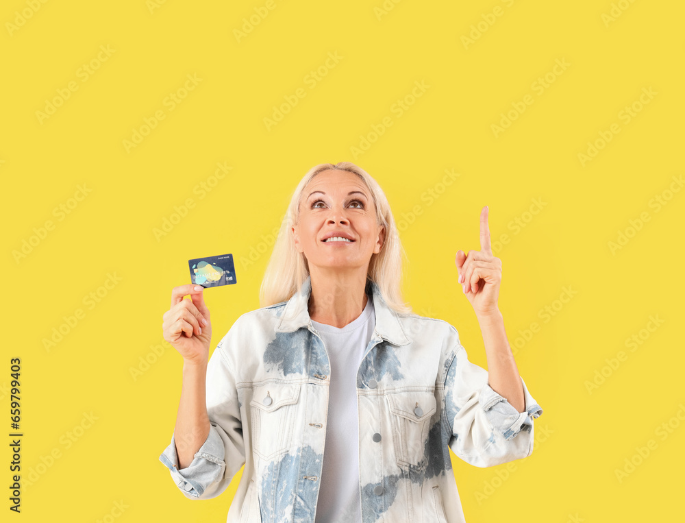 Mature woman with credit card pointing at something on yellow background