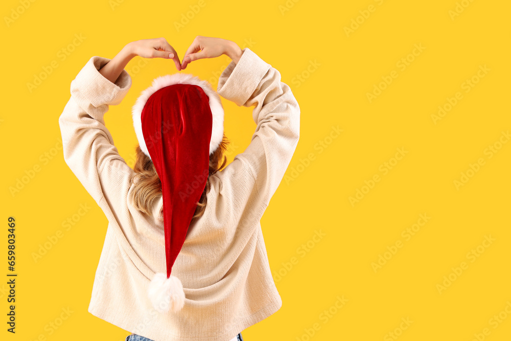 Young woman in Santa hat making heart gesture on yellow background, back view