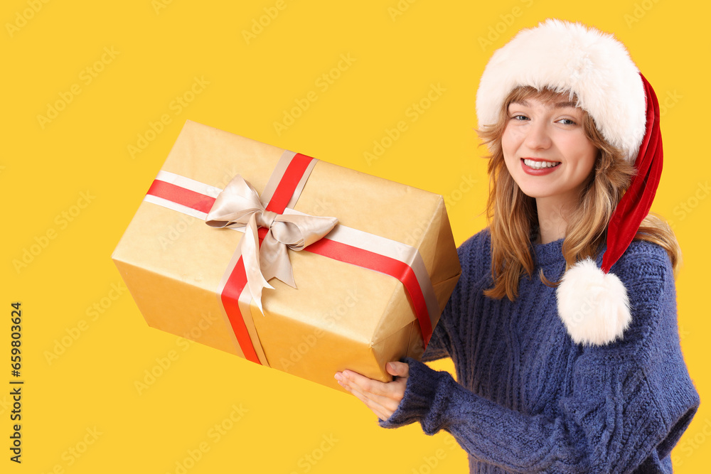 Young woman in Santa hat with Christmas gift on yellow background