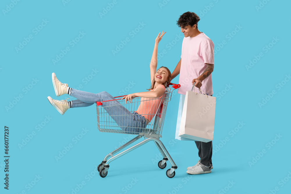 Young man with bags and his girlfriend in shopping cart on blue background