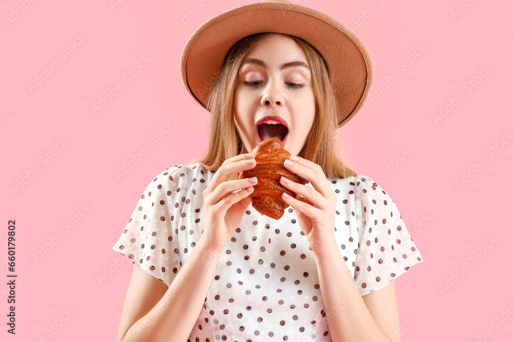 Beautiful young woman in wicker hat eating tasty croissant on pink background