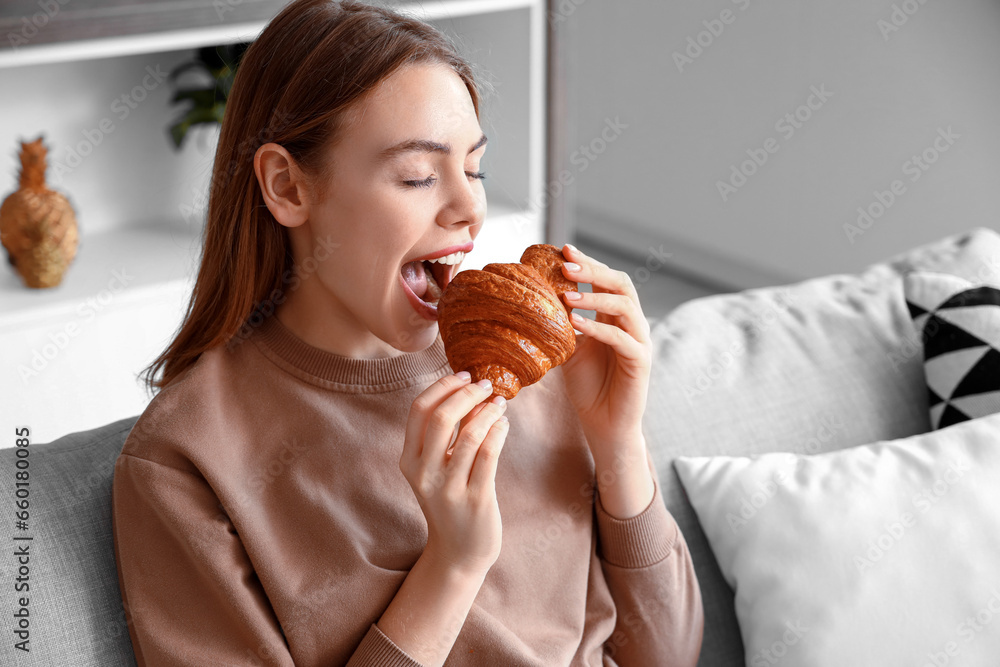 Beautiful young woman eating tasty croissant and sitting on sofa in living room