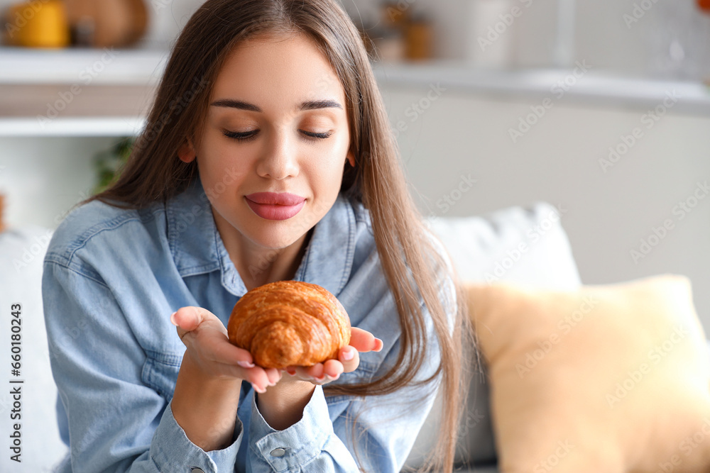 Beautiful young woman with tasty croissant sitting on sofa in living room