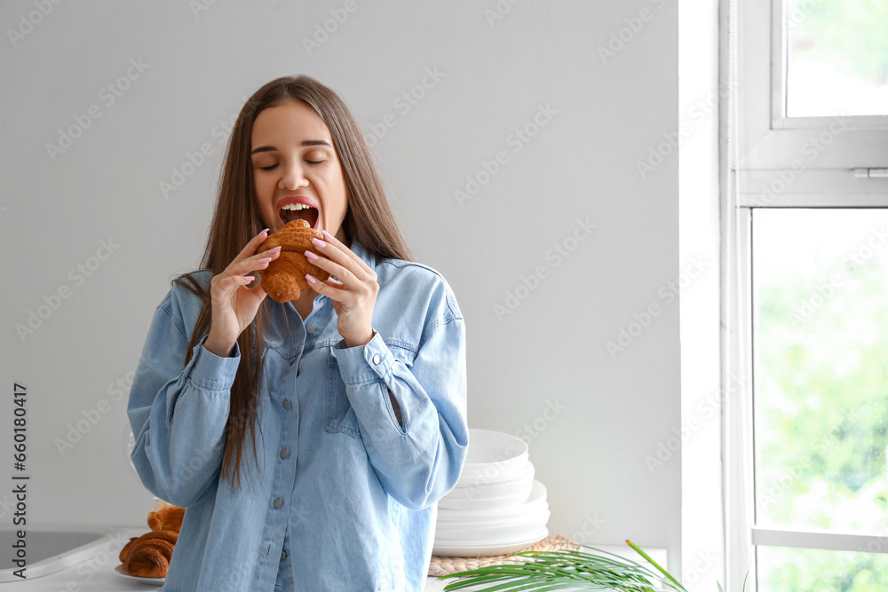 Beautiful young woman eating tasty croissant in kitchen