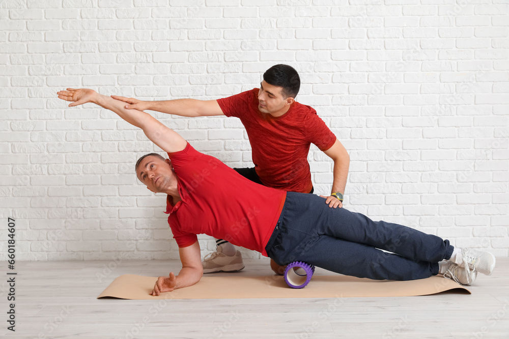 Mature man training with foam roller and rehabilitation therapist near white brick wall