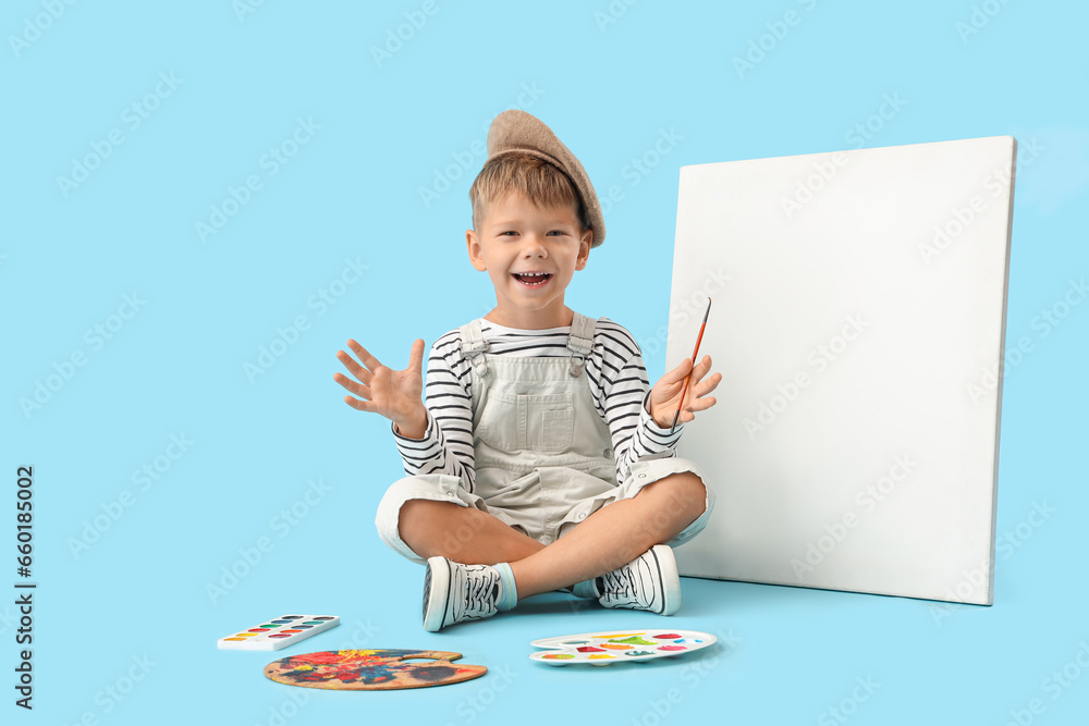 Cute little artist with paint palettes and canvas sitting on blue background