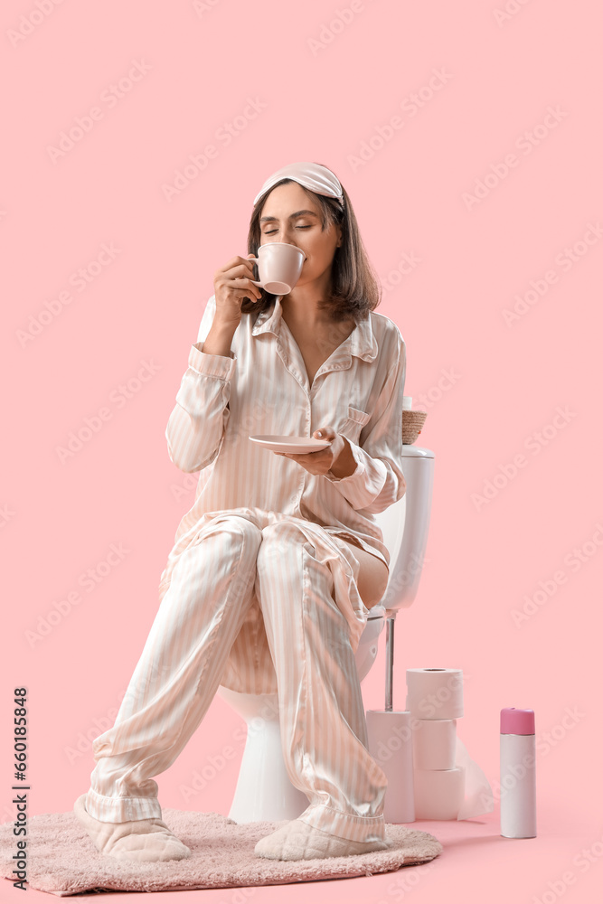 Young woman in pajamas drinking coffee on toilet bowl against pink background